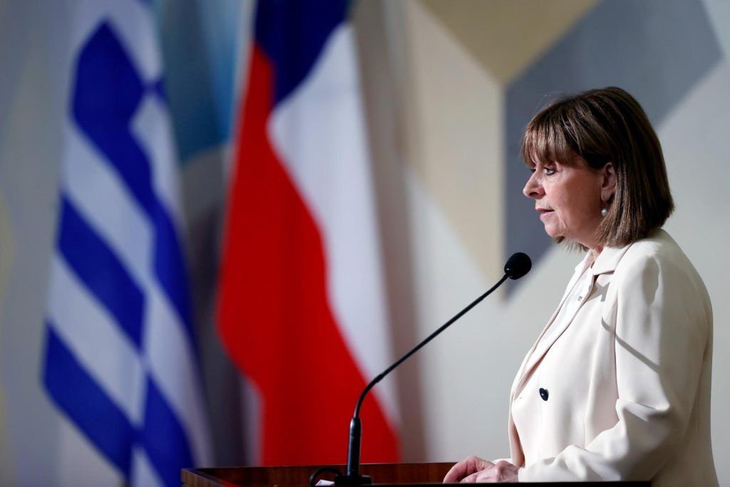 En la foto aparece la actual presidenta de Grecia, Katerina Sakellaropoulou. En la imagen, la mandataria aparece de perfil, hablando al micrófono. Viste con blazer blanco y de fondo se ven las banderas de Grecia y Chile, respectivamente.