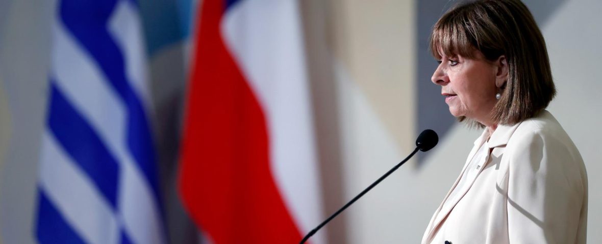En la foto aparece la actual presidenta de Grecia, Katerina Sakellaropoulou. En la imagen, la mandataria aparece de perfil, hablando al micrófono. Viste con blazer blanco y de fondo se ven las banderas de Grecia y Chile, respectivamente.