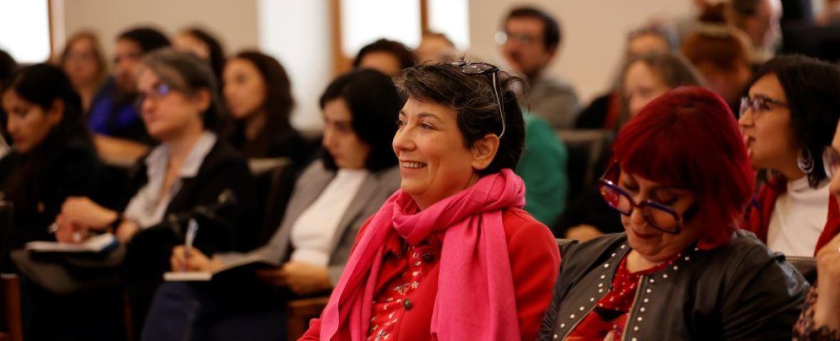 En la foto aparece el público que asistió a la charla. En ella, se observa a un grupo de mujeres sentada mirando hacia la testera. El foco de la cámara está sobre una mujer de pelo corto, vestida de chaleco y bufanda fucsia, que sonríe. A su lado, una mujer de anteojos rojos toma apuntes.